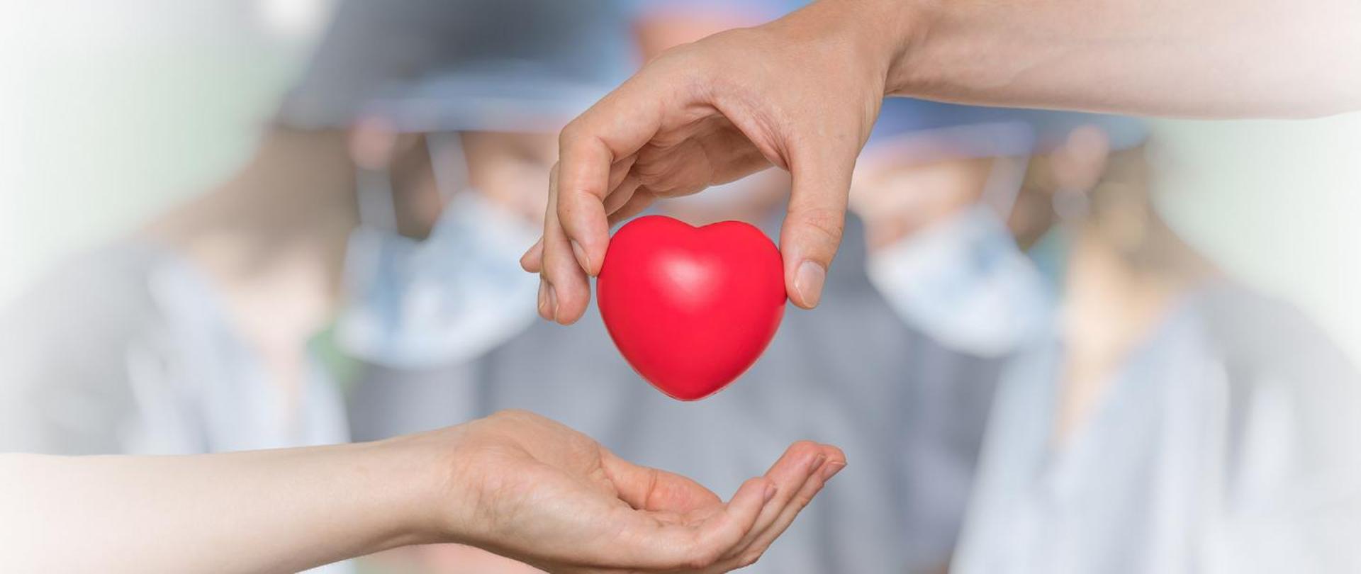Grafika oparta na fotografii gdzie z dłoni do dłoni przekazywane jest plastikowe serduszko, a w tle rozmyte postacie lekarzy podczas prawdopodobnie operacji transplantacji serca.