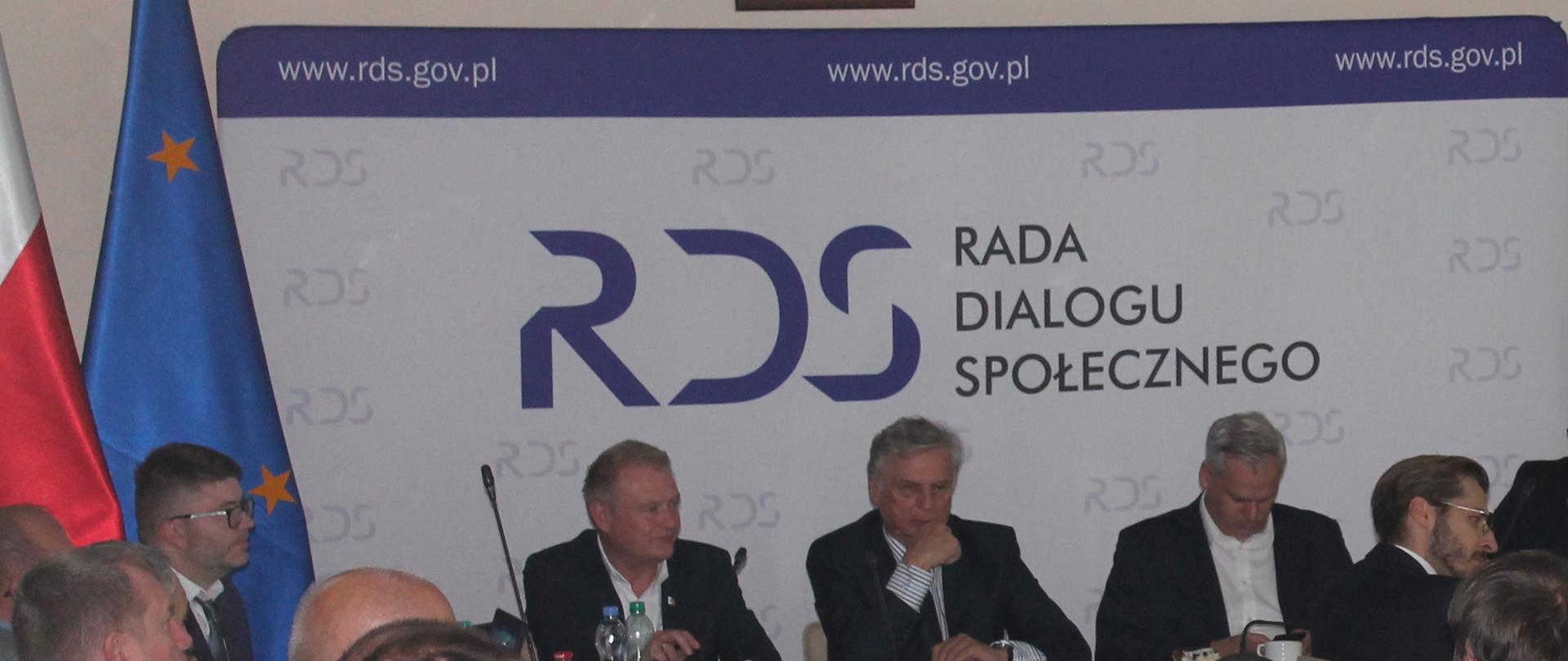 Zdjęcie przedstawia stół prezydialny za którym siedzą: Karol Rabenda - Podsekretarz Stanu w Ministerstwie Aktywów Państwowych, Współprzewodniczący Zespołów ze strony rządowej, Pan Krzysztof Kisielewski, Współprzewodniczący Zespołu Trójstronnego ds. Branży Energetycznej ze strony związków zawodowych, Pan Ryszard Wasiłek, Współprzewodniczący Zespołu Trójstronnego ds. Branży Energetycznej ze strony pracodawców. W tle siedzących znajduje się baner z napisem Centrum Partnerstwa Społecznego Dialog im. Andrzeja Bączkowskiego. Po lewej stronie zdjęcia stoją dwie flagi: Polska i UE. 