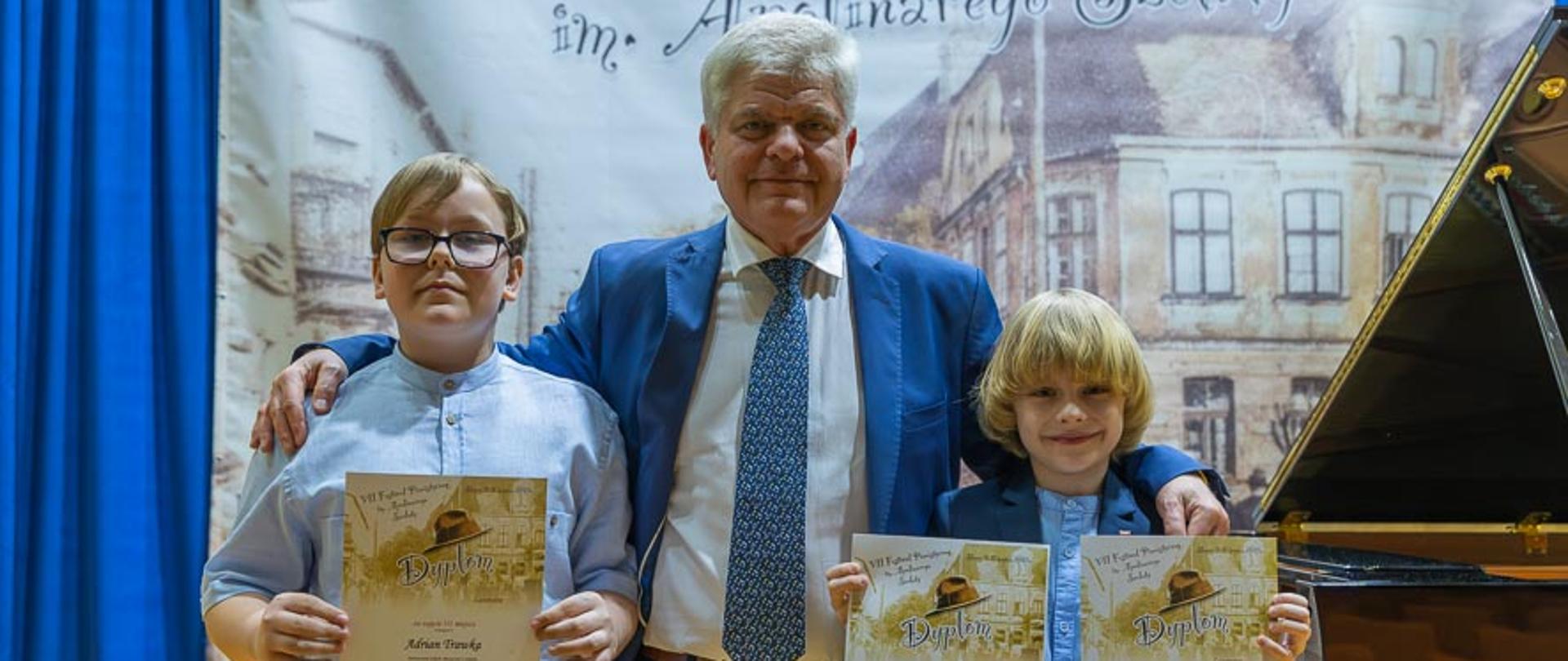 Dwóch chłopców trzymających dyplomy, pomiędzy nimi mężczyzna, obok fortepian, z animi baner festiwalowy