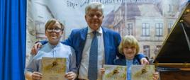 Dwóch chłopców trzymających dyplomy, pomiędzy nimi mężczyzna, obok fortepian, z animi baner festiwalowy