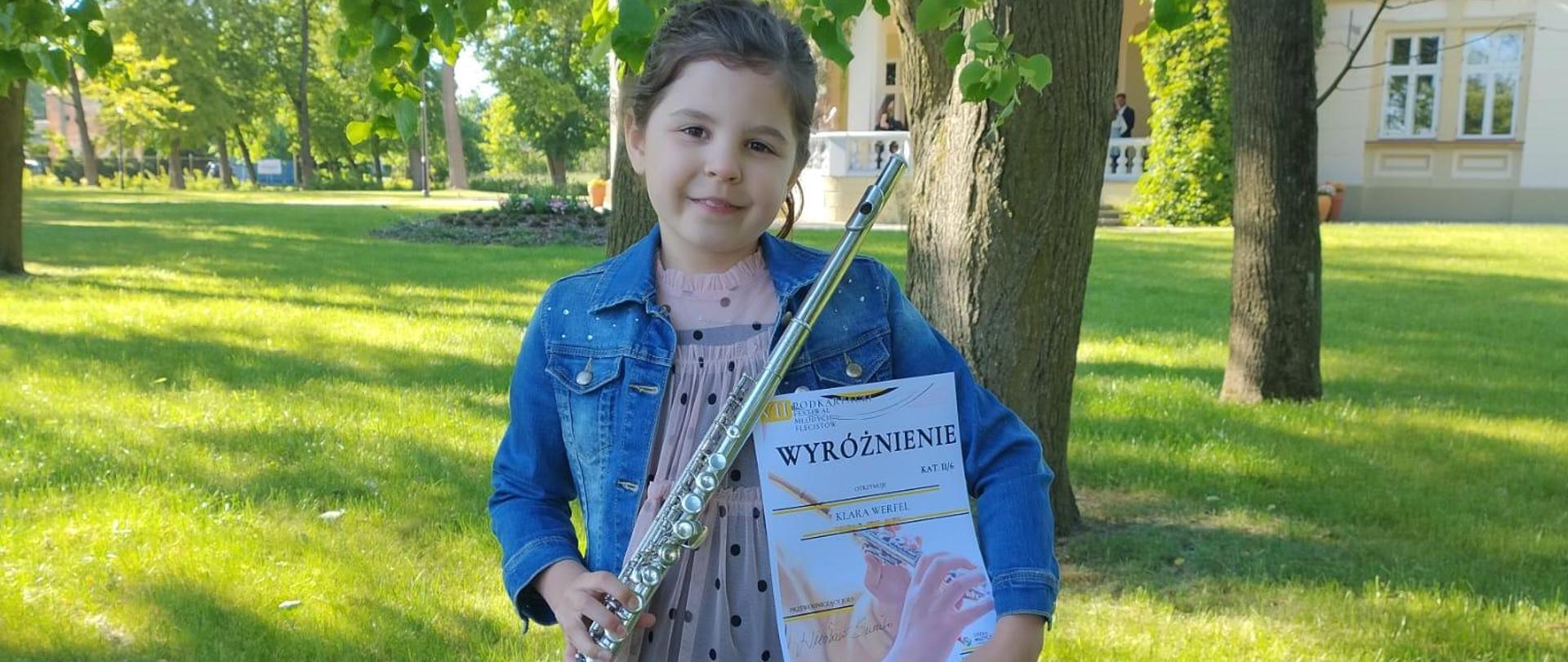 Uczennica trzymająca dyplom laureata oraz flet. Z tyłu widoczne liściaste drzewa oraz fragment budynku szkoły.