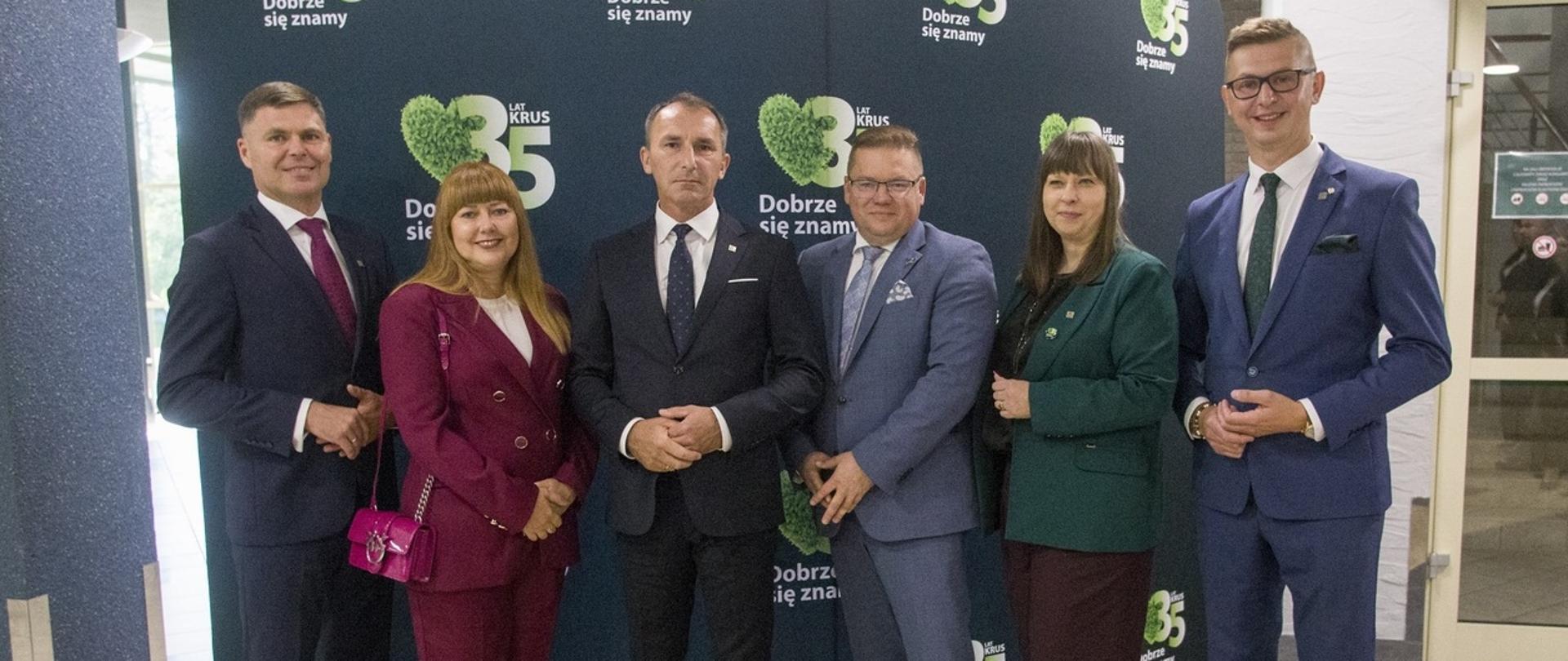 Dwie kobiety i czterech mężczyzn pozuje do zdjęcia na tle ścianki 35-lecia KRUS. Prezes stoi w środku grupy. Wszyscy uśmiechają się do zdjęcia.