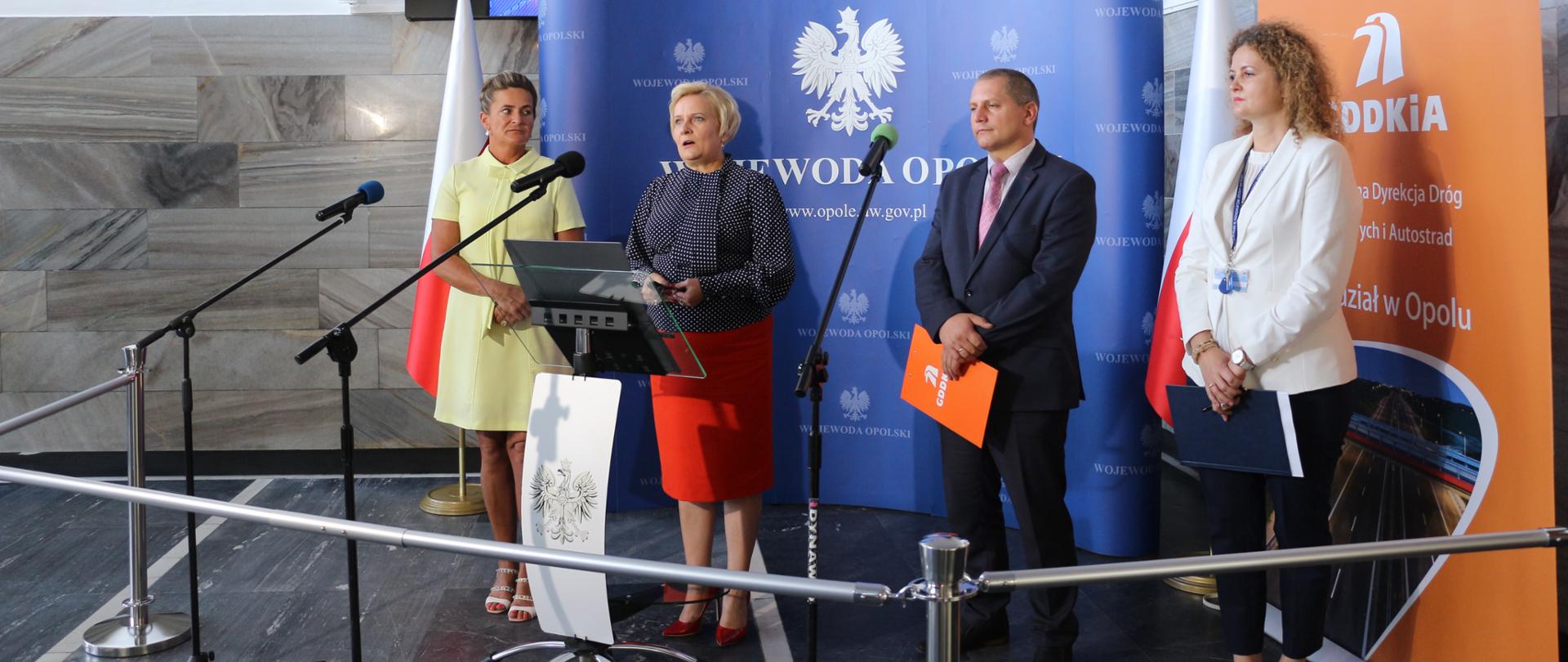 Na zdjęciu 4 osoby. Od lewej: ubrana w żółtą sukienkę poseł Czochara, wicewojewoda opolski ubrana w czerwoną spódnicę i granatową koszulę, dyrektor opolskiego oddziału GDDKiA ubrany w garnitur oraz dyrektor wydziału infrastruktury urzędu, która ubrana jest w biały żakiet.
Wszyscy stoją za mikrofonami, które na statywach. Dodatkowo, przed wicewojewodą stoi mównica, na którą postawiono laptopa. W tle granatowa ścianka z logo Wojewody oraz monitor - duży ekran, na którym wyświetlona jest prezentacja dot. inwestycji drogowych. N pierwszym planie, nieco od dołu, widać pachołki odgradzające dziennikarzy od mówców.
