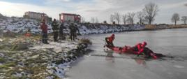 Zdjęcie przedstawia strażaków ćwiczących na lodzie na zbiorniku. Trzech znajduje się na tafli lodu na saniach lodowych, pięcioro asekuruje ich z brzegu przy użyciu linek. W tle widać samochody bojowe straży na brzegu zbiornika