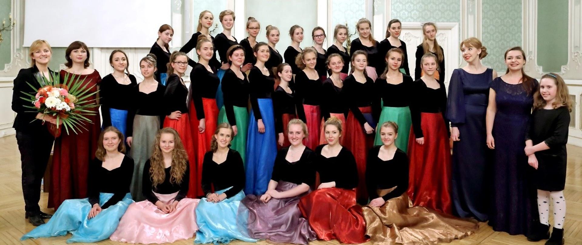 Dziewczęta stojące w trzech rzędach ubrane w czarne bluzki i długie spódnice o różnych jednolitych kolorach. Pierwszy rząd przykucnięty. Z lewej strony stoi dyrygentka z wiązanką kwiatów.