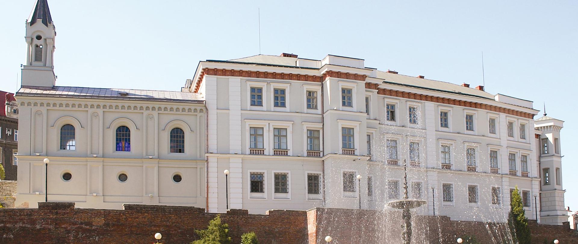 Jasny, klasycystyczny budynek z wysoką wieżą po lewej stronie, stojący na podwyższeniu z ceglaną murawą. Na pierwszym planie fontanna tryskająca wodą, wokół zieleń i latarnie, nad całością bezchmurne niebo.
