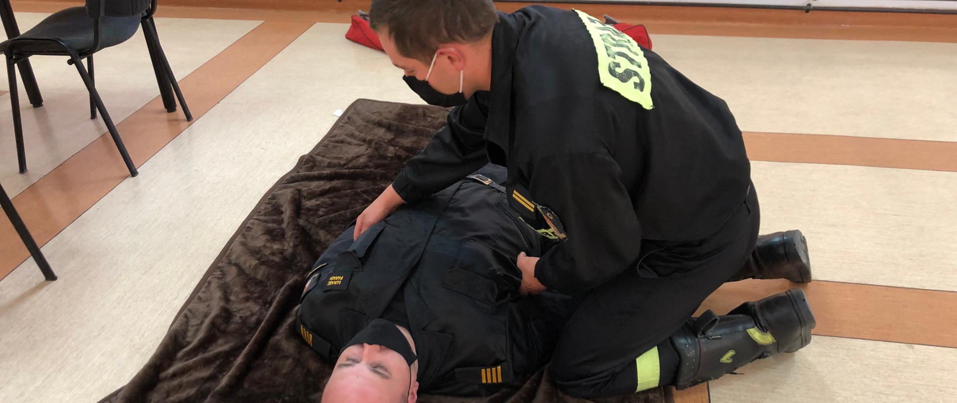 Na zdjęciu strażak-ratownik wykonuje symulację badania urazowego osoby poszkodowanej. Badanie rozpoczyna od głowy, następnie bada szyję, obojczyki, klatkę piersiową, brzuch, miednicę, kończyny dolne, kończyny górne oraz plecy i pośladki. Badanie urazowe polega na szybkim zidentyfikowaniu krwawień oraz innych nieprawidłowości poszczególnych części ciała.