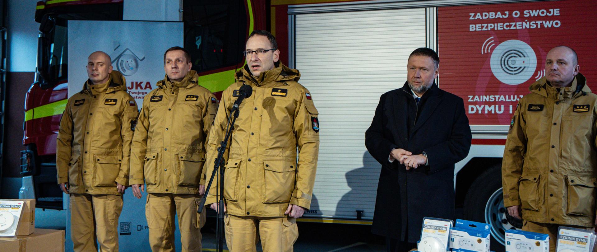 Zdjęcie przedstawia konferencję prasową odbywającą się w garażu jednostki straży pożarnej, na tle nowoczesnego wozu strażackiego. W centrum stoi mężczyzna w okularach i piaskowym mundurze służbowym Państwowej Straży Pożarnej, który przemawia do mikrofonu ustawionego na statywie. Po jego lewej stronie stoją dwaj inni strażacy w identycznych mundurach, a po prawej stronie – jeden strażak oraz mężczyzna w ciemnym płaszczu i garniturze.