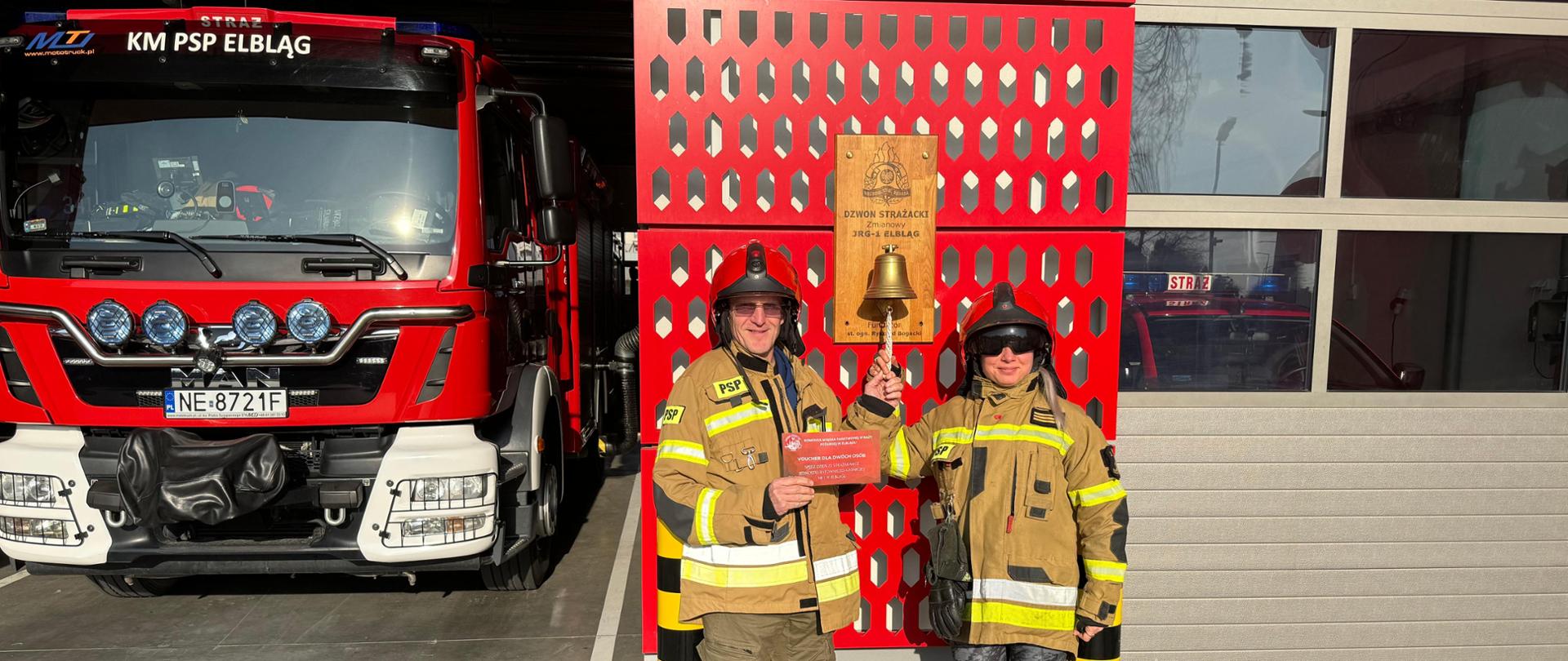 Przed remizą obok strażackiego dzwonka stoi małżeństwo ubrane w strażackie mundury i hełmy. Mężczyzna w ręku trzyma czerwony kartonik z napisem voucher. Po lewej stronie widać pojazd pożarniczy znajdujący się w garażu.
