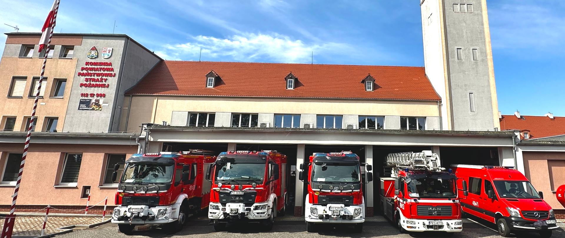 Strażnica KP PSP Kościan. Na zdjęciu pięć samochodów pożarniczych, które częściowo wyjechały z garażu i stoją w jednym rzędzie. Za nimi strażnica Komendy Powiatowej PSP w Kościanie. Po lewej stronie masz z biało-czerwoną flagą.