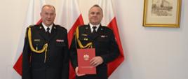 Dwóch funkcjonariuszy w mundurach wyjściowych ze sznurem stoi obok siebie jeden z nich trzyma czerwoną teczkę za nimi znajdują się trzy flagi Polski na ścianie wisi obraz.