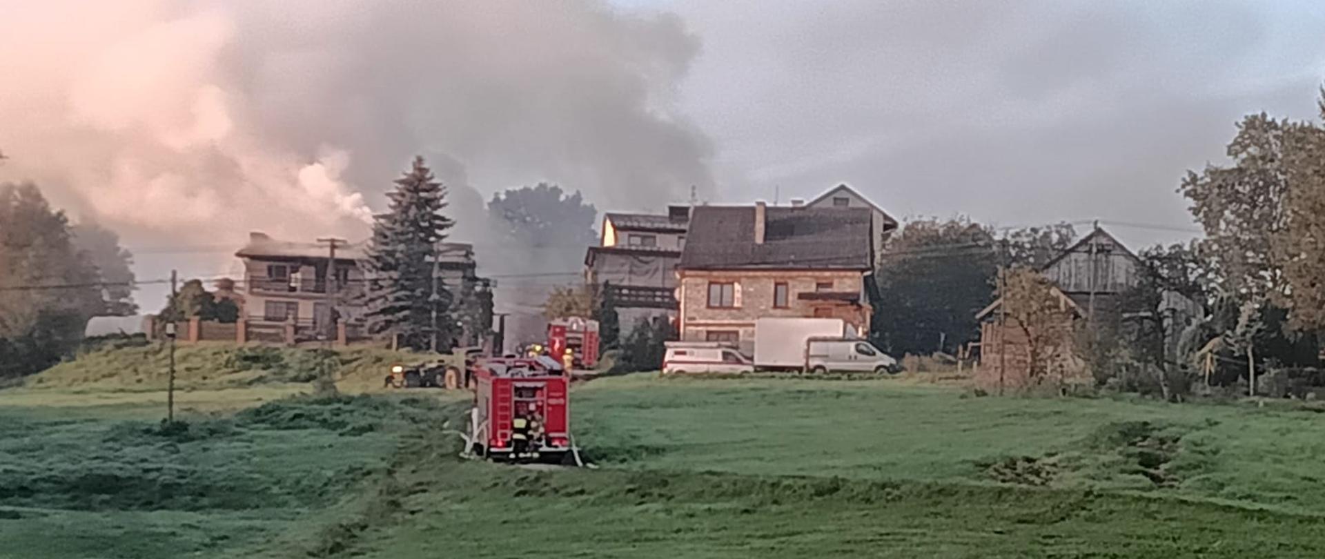 Zdjęcie z miejsca zdarzenia godzinny poranne. Widoczny teren wiejski z zabudowaniami mieszkalnymi i gospodarczymi. W centralnej części kadru, na drodze dojazdowej do zabudowań widoczne są zaparkowane samochody ratowniczo-gaśnicze przy których widać rozwinięte węże strażackie. Nad kompleksem budynków unoszą się kłęby jasnego dymu, co świadczy o trwającym pożarze. 