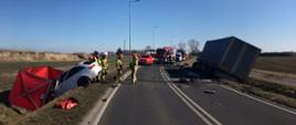 Samochody strażackie i osobowe stoją na jezdni. Cięzarówka stoi wywrucona w rowie. 