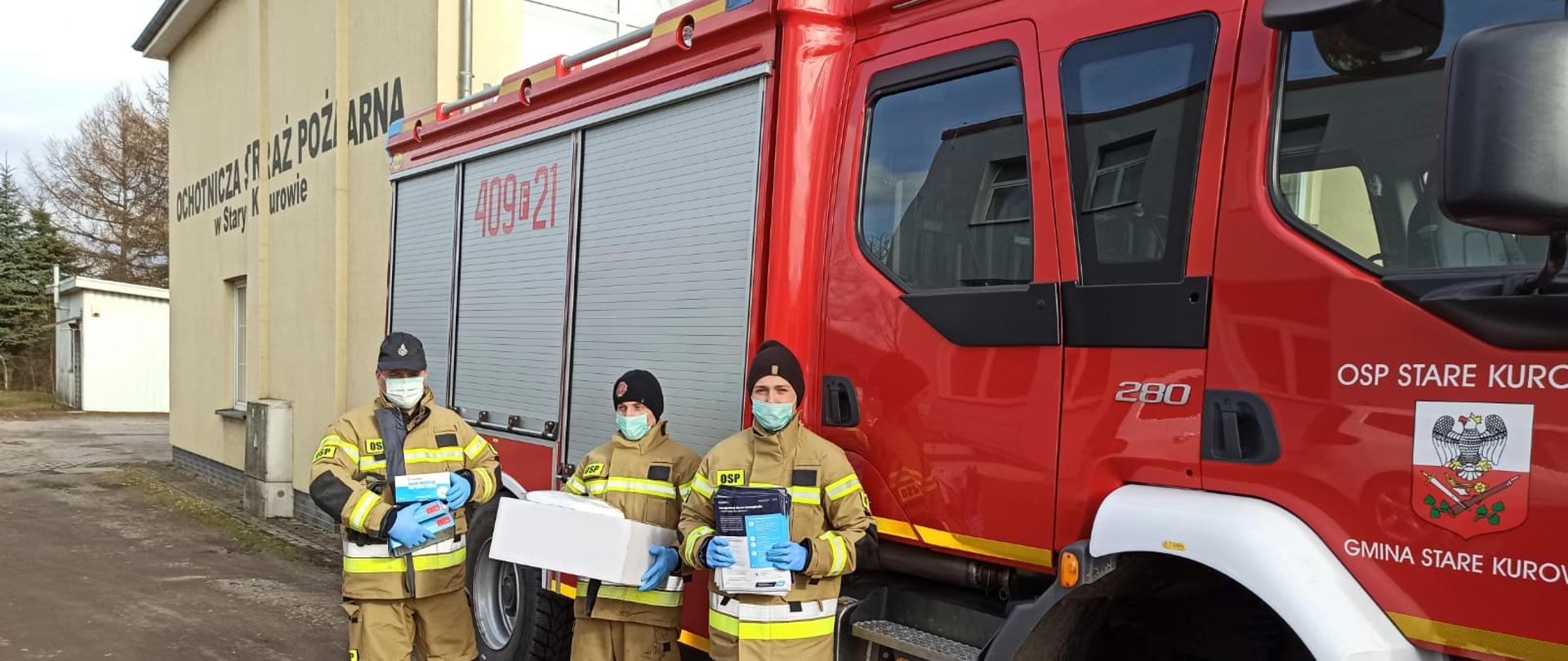 Fotografia przedstawia ujęcie prawego boku samochodu gaśniczego. Przed samochodem stoi trzech strażaków OSP w ubraniach specjalnych, maseczkach i rękawicach nitrylowych, trzymają ulotki informacyjne i maseczki przeznaczone do kolportażu. Samochód stoi przed remizą OSP Stare Kurowo.