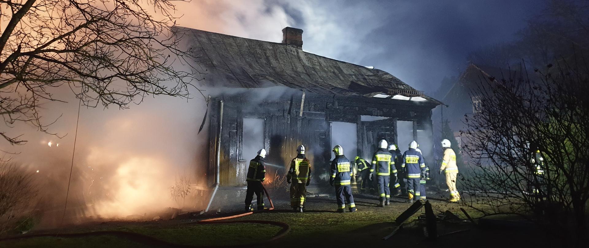 Zdjęcie spalonego domu drewnianego, przed którym stoją strażacy