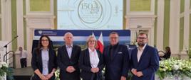 Grupa ludzi w strojach biznesowych stojących na tle ekranu z logo jubileuszu 150-lecia i banera Ministerstwa Rolnictwa i Rozwoju Wsi