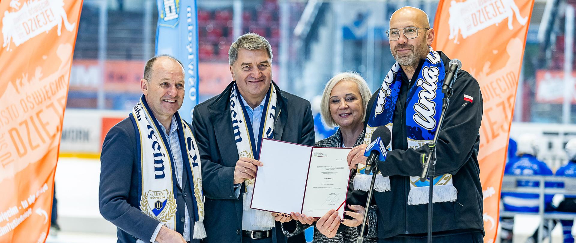 Konferencja prasowa. Minister sportu i turystyki Jakub Rutnicki i trzy inne osoby (1 kobieta i 2 mężczyzn) stoją za mikrofonami i prezentują decyzję o dofinansowaniu rozbudowy hali lodowej. W tle widać lodowisko.