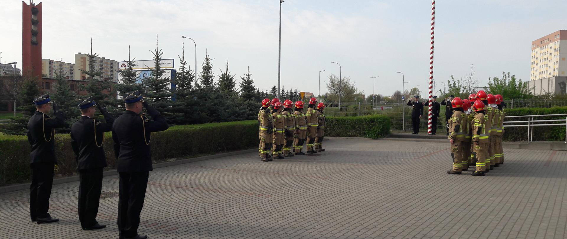 Zdjęcie przedstawia strażaków stojących na placu przed budynkiem straży pożarnej podczas uroczystego wciągnięcia flagi na masz. 