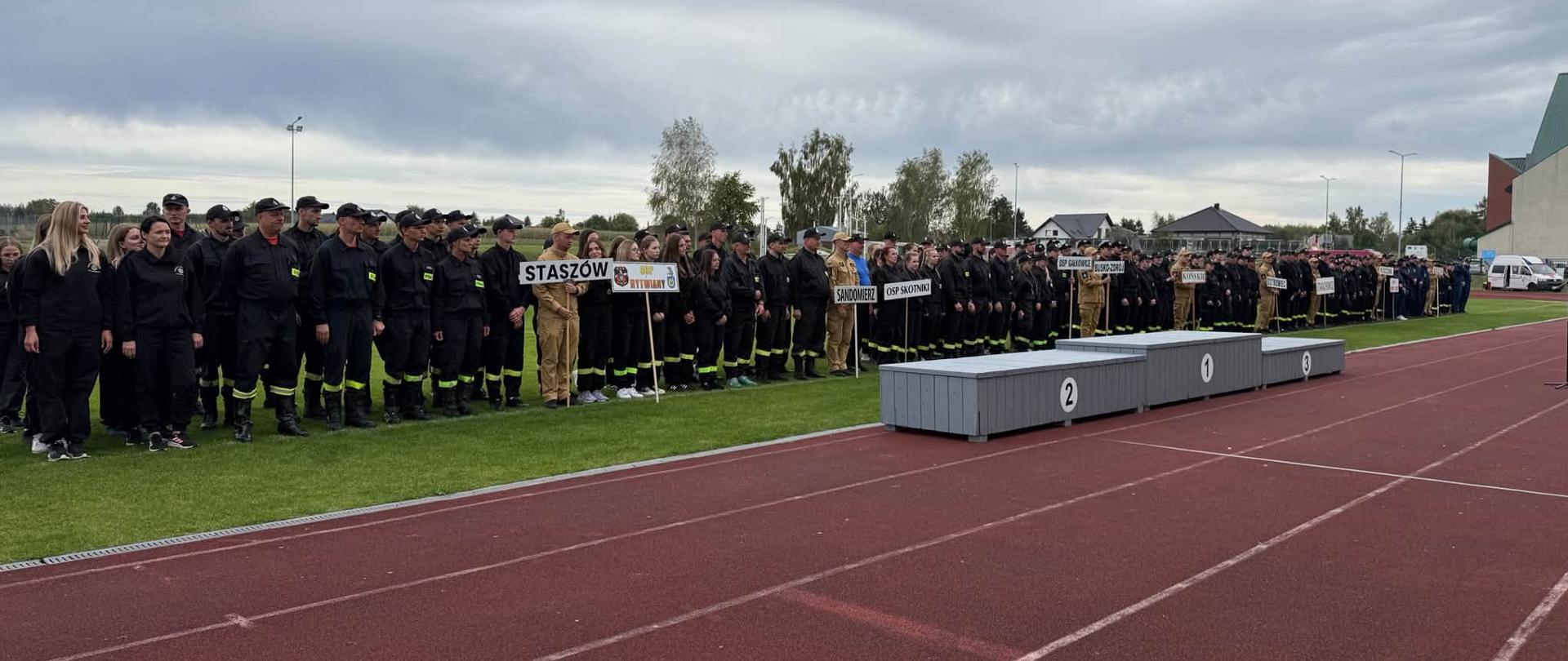 Fotografia przedstawia uczestników zawodów sportowych po zakończeniu rywalizacji stojących za podium dla zwycięzców ustawionym na bieżni.