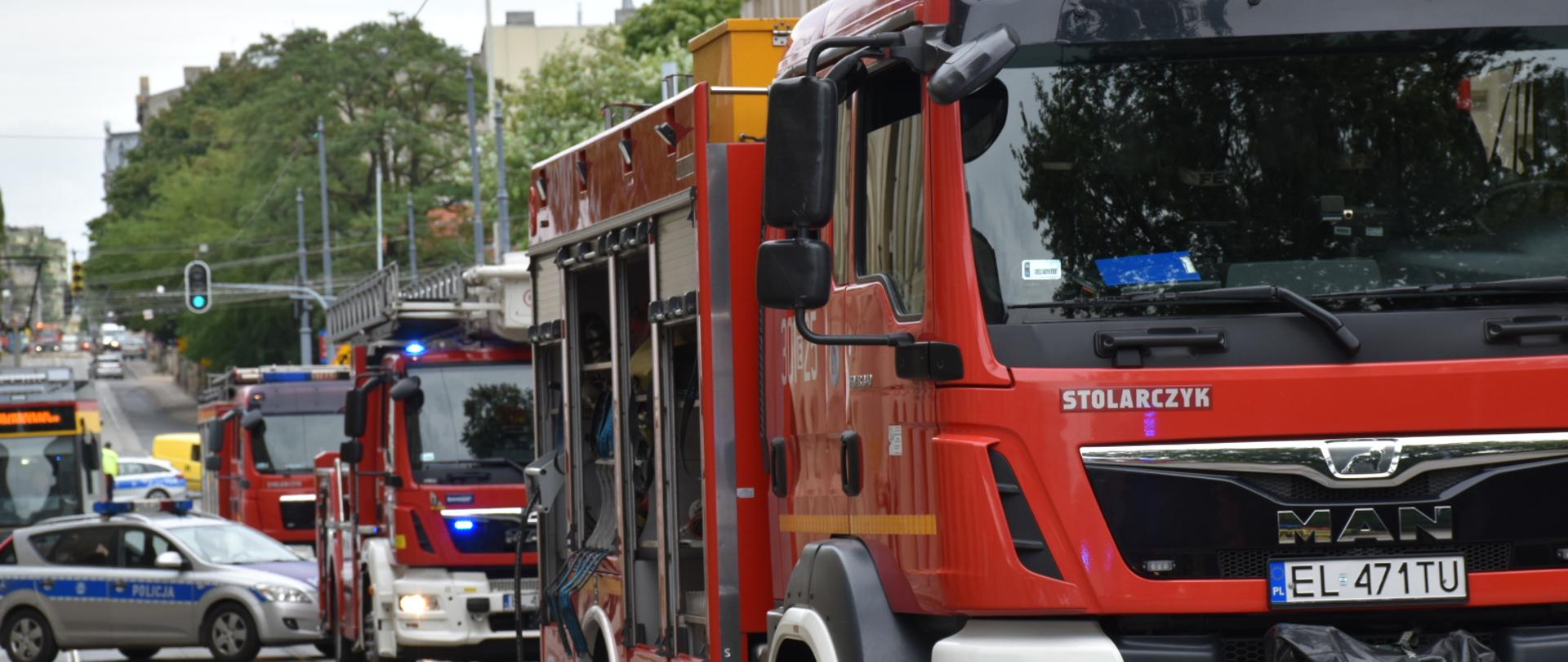 Samochody pożarnicze stojące na ulicy, w tle radiowóz policyjny