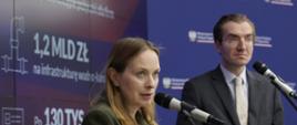 
Minister Katarzyna Pełczyńska-Nałęcz and Deputy Minister Jan Szyszko stand on stage in front of microphones