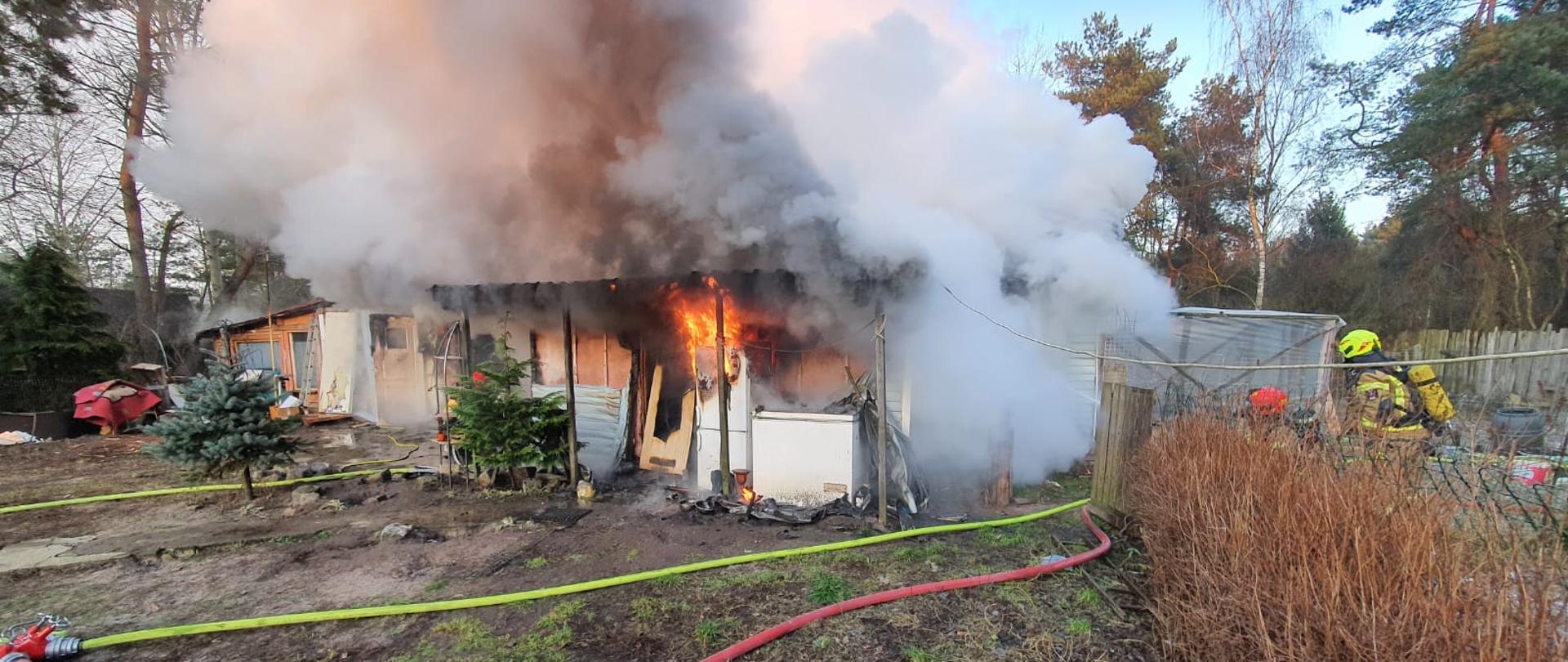 pożar budynku parterowego typu ogrodowego, płomienie pod płaskim dachem, duże zadymienie, rozwinięte węże strażackie (czerwony linia główna, rozdzielacz, żółty dwie linie gaśnicze po dwóch stronach budynku) na terenie działki, kępy zielonej trawy, 