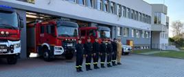 Grupa strażaków w ubraniach koszarowych przed samochodami pożarniczymi z włączonymi sygnałami świetlnymi niebieskimi na placu zewnętrznym KPPSP W Opatowie