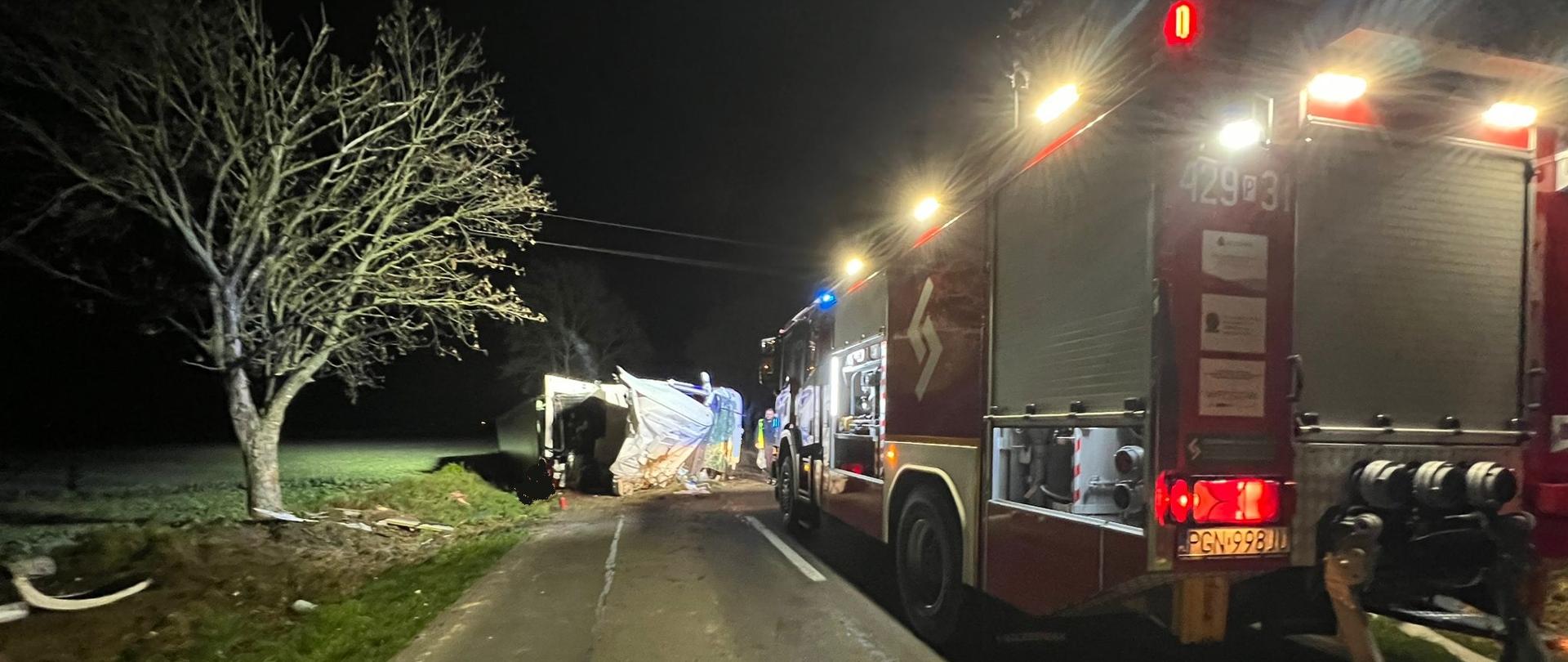 Na zdjęciu widać samochód pożarniczy oświetlający teren akcji. Przed samochodem pożarniczym leży na boku samochód ciężarowy, który tarasuje przejazd DW190