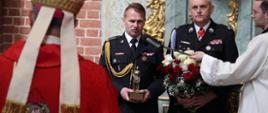 Oficer PSP i strażak OSP w mundurach galowych, stoją przed biskupem i wręczają upominki w podziękowaniu za eucharystię. Strażak z prawej strony trzyma kwiaty biało-czerwone, strażak z lewej statuetkę świętego 
