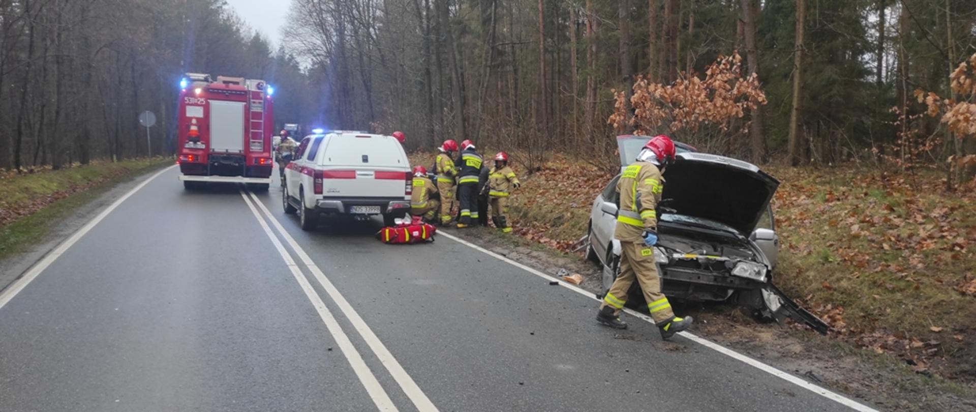 Rozbity samochód osobowy stoi w rowie. Strażacy udzielają pomocy poszkodowanym. 
