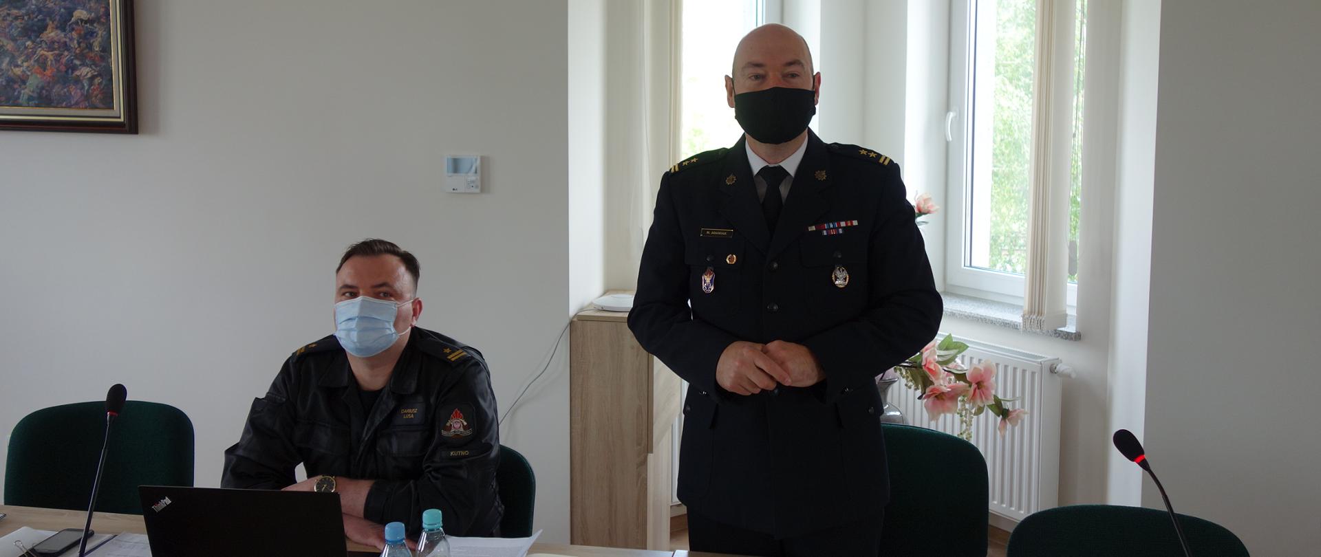 Stojąc przemawia Zastępca Komendanta Państwowej Straży Pożarnej w mundurze wyjściowym i czarną maseczką ochronną na ustach i nosie. Na stole widać notes, okulary, wodę butelkowaną, dokumenty i laptopa przy którym siedzi Naczelnik Wydziału ds. Operacyjno-Szkoleniowych.