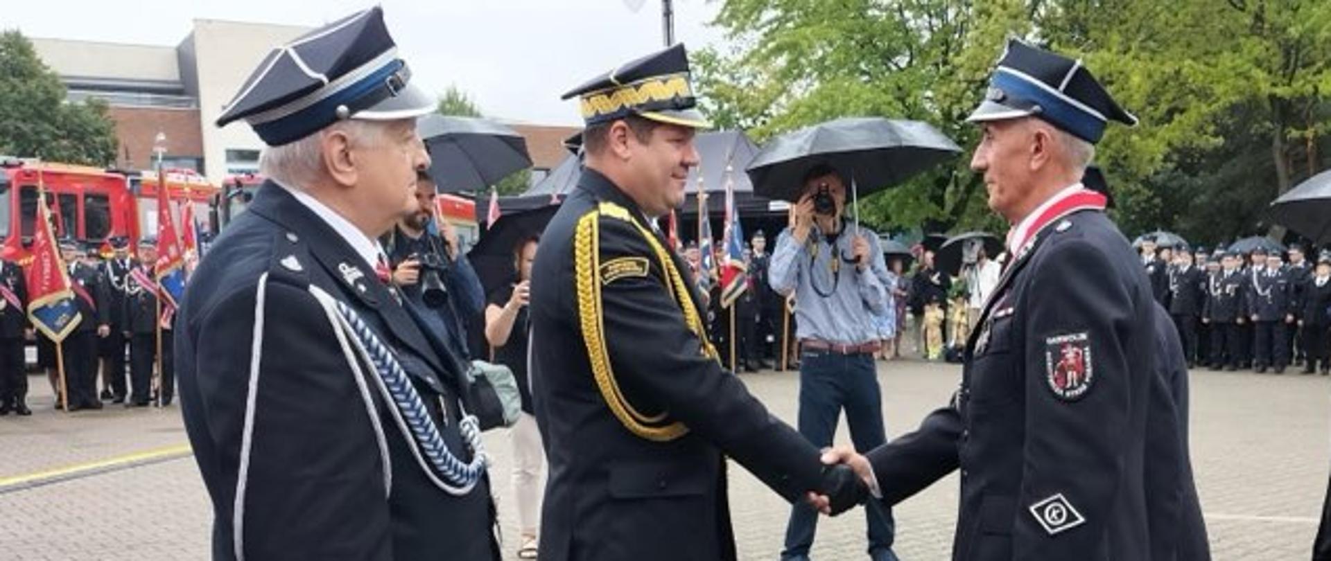 150-lecie Ochotniczej Straży Pożarnej w Garwolinie. Dwóch strażaków w mundurach stojąc na przeciwko siebie uściskuje sobie prawe dłonie. Obok nich stoi inny strażak w mundurze. W tle widoczne są samochody strażackie, sztandary i zgromadzeniu ludzie. 