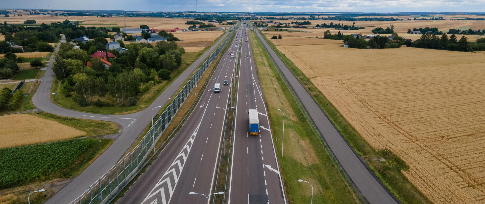 Widok z lotu ptaka na drogę ekspresową S12/S17 między Lublinem a Piaskami, przebiegającą przez pola uprawne; po jezdniach poruszają się samochody i ciężarówka, nad trasą wiszą ciemne chmury. 