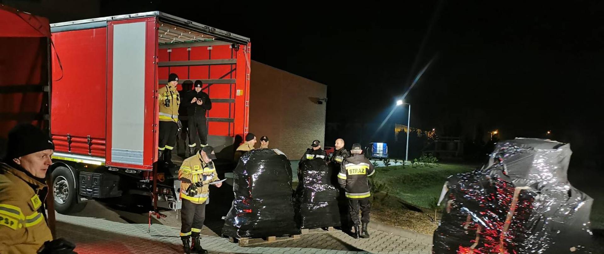 wieczór, na sużym placu strażacy pakuja do samochodu duże palety owinięte czarnym streczem