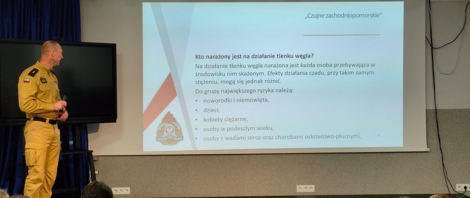 Strażak w żółtym mundurze stoi na scenie obok ekranu z prezentacją o tlenku węgla. Przed nim siedzi grupa uczniów, którzy uważnie słuchają. Na slajdzie widoczny jest tekst wyjaśniający, na czym polega działanie tlenku węgla i dlaczego jest groźny.
