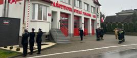Zdjęcie przedstawia grupę strażaków stojących na baczność i salutujących na brukowanym placu przed budynkiem.
W pierwszym planie, po lewej stronie, stoi strażak w jasnobeżowym mundurze polowym (z napisem "PAŃSTWOWA STRAŻ POŻARNA" na plecach), zwrócony tyłem do aparatu i salutujący. Przed nim, lekko z prawej, stoi szereg dziewięciu innych strażaków w ciemnych (czarnych) ubraniach koszarowych, również stojących na baczność i salutujących.
Po prawej stronie zdjęcia widoczny jest fragment wejścia do budynku komendy Straży Pożarnej z czerwonymi akcentami i schodami. Na placu w głębi, po prawej, znajduje się maszt z częściowo widoczną polską flagą (biało-czerwona). W oddali, bliżej wejścia do budynku, stoi jedna postać w ciemnym mundurze, również salutująca.
Tło stanowią domy mieszkalne za ciemnym ogrodzeniem, typowe dla zabudowy podmiejskiej lub małego miasteczka. Dzień jest pochmurny, a bruk jest mokry od deszczu.