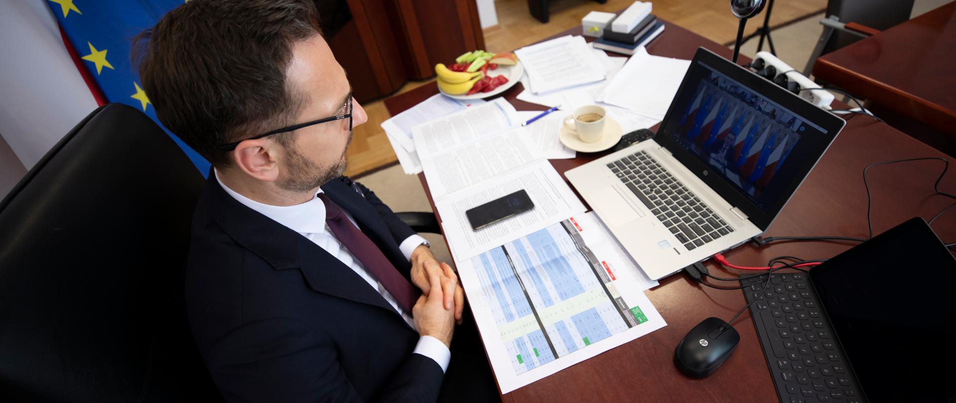 Wiceminister Waldemar Buda siedzi w gabinecie przy biurku. Na blacie laptop, dokumenty, filiżanka z kawą i talerz z owocami.