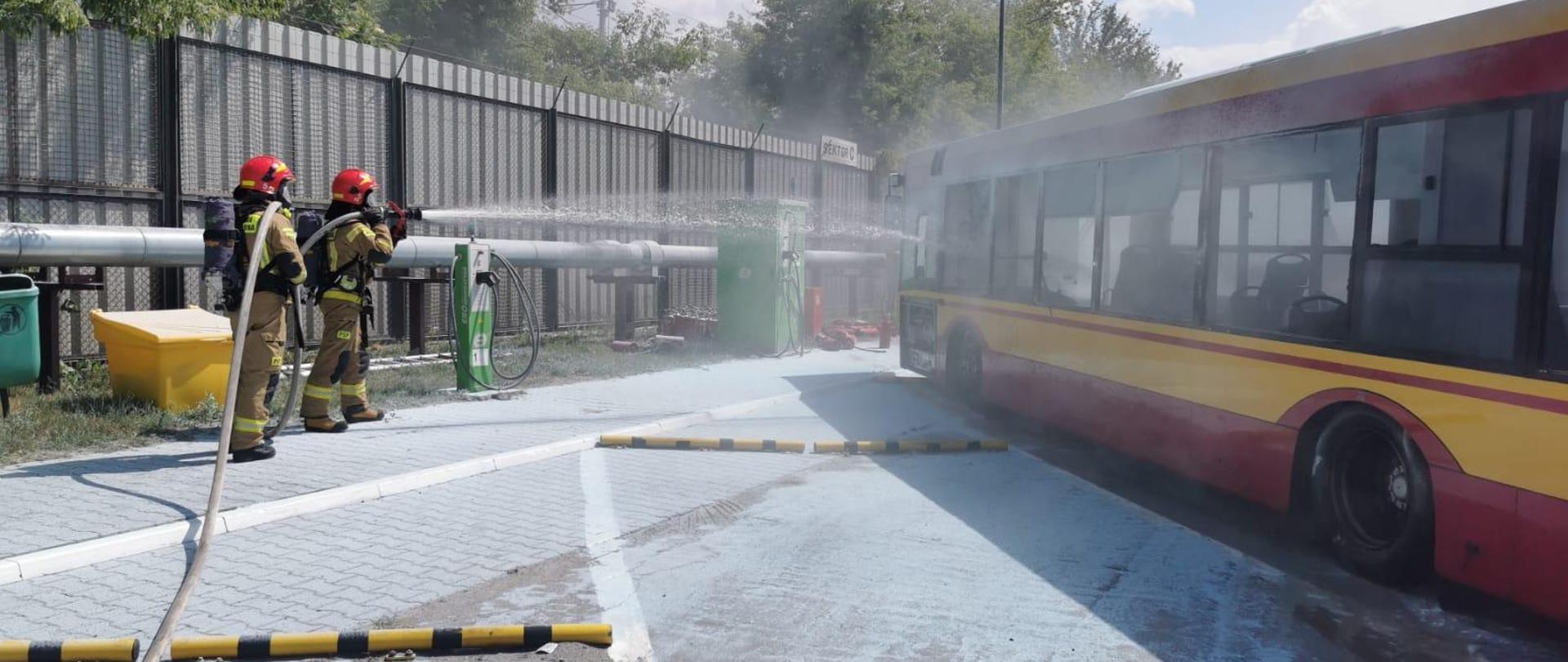 Funkcjonariusze podczas ćwiczeń, w ubraniu specjalnym, podają prąd wody na autobus.