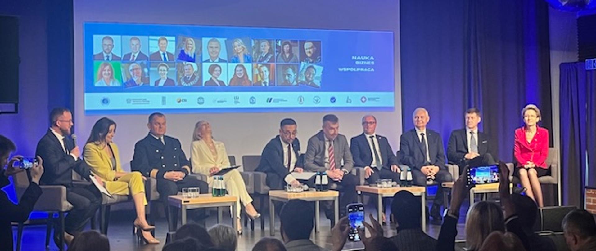 Na podium siedzą uczestnicy panelu dyskusyjnego – rektorzy uczelni wyższych oraz minister nauki i szkolnictwa wyższego. W tle widać logo konferencji i ekran z prezentacją. Paneliści prowadzą rozmowę na temat szkolnictwa wyższego.