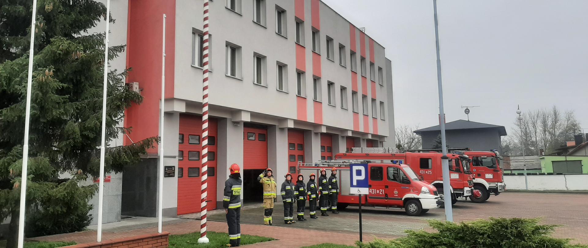 Na zdjęciu widać strażaków stojących w szeregu przed budynkiem komendy powiatowej, oddających honory podczas wciągania na maszt flagi państwowej 