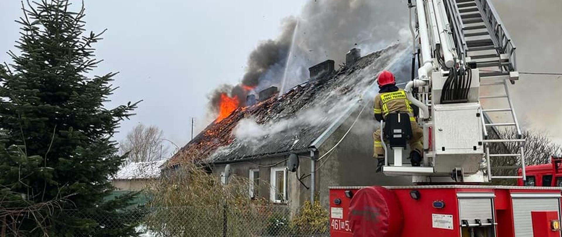Strażacy gaszą pożar dachu w budynku mieszkalnym, przy wykorzystaniu podnośnika hydraulicznego. 