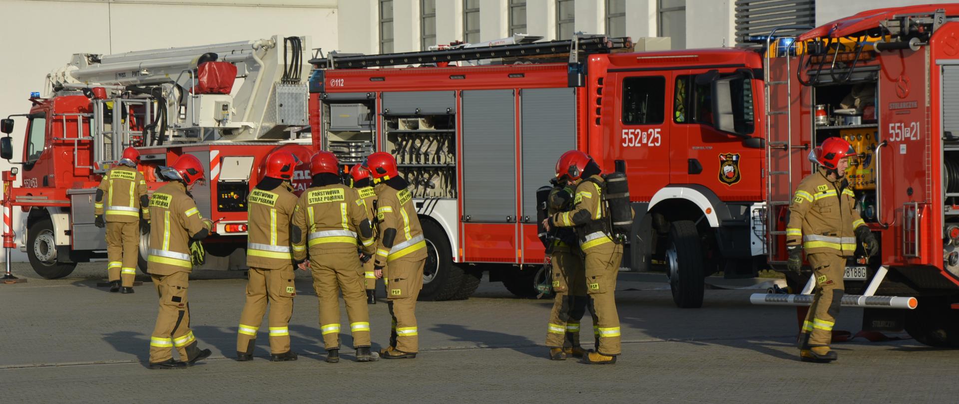 Na zdjęciu widać dwa samochody gaśnicze, drabinę mechaniczną oraz strażaków przygotowujących się do ćwiczeń.