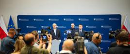 Minister Tomasz Siemoniak (po lewej), minister Marcin Kierwiński (po prawej) oraz komendant główny Policji gen. insp. Marek Boroń (w środku) stoją na tle niebieskiej ścianki ze zmuliplikowanym logo MSWiA (orzeł, napis Ministerstwo Spraw Wewnętrznych i Administracji oraz biało-czerwony pasek). Ministrowie stoją za szarymi mównicami z czarnymi mikrofonami kierunkowymi. Po prawej stronie ścianki biało-czerwona flaga Polski, a po lewej stronie ścianki flaga UE (niebieska z żółtymi gwiazdkami) oraz biało-czerwona Polski. Przed mównicami (na pierwszym planie) stoją operatorzy, fotoreporterzy i dziennikarze z kamerami i aparatami fotograficznymi.