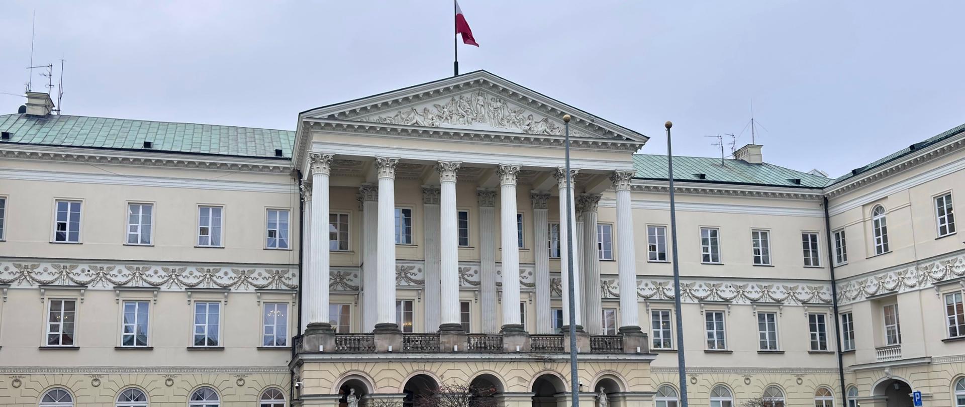 Biały duży budynek, z kolumnami, flaga Polski na dachu.