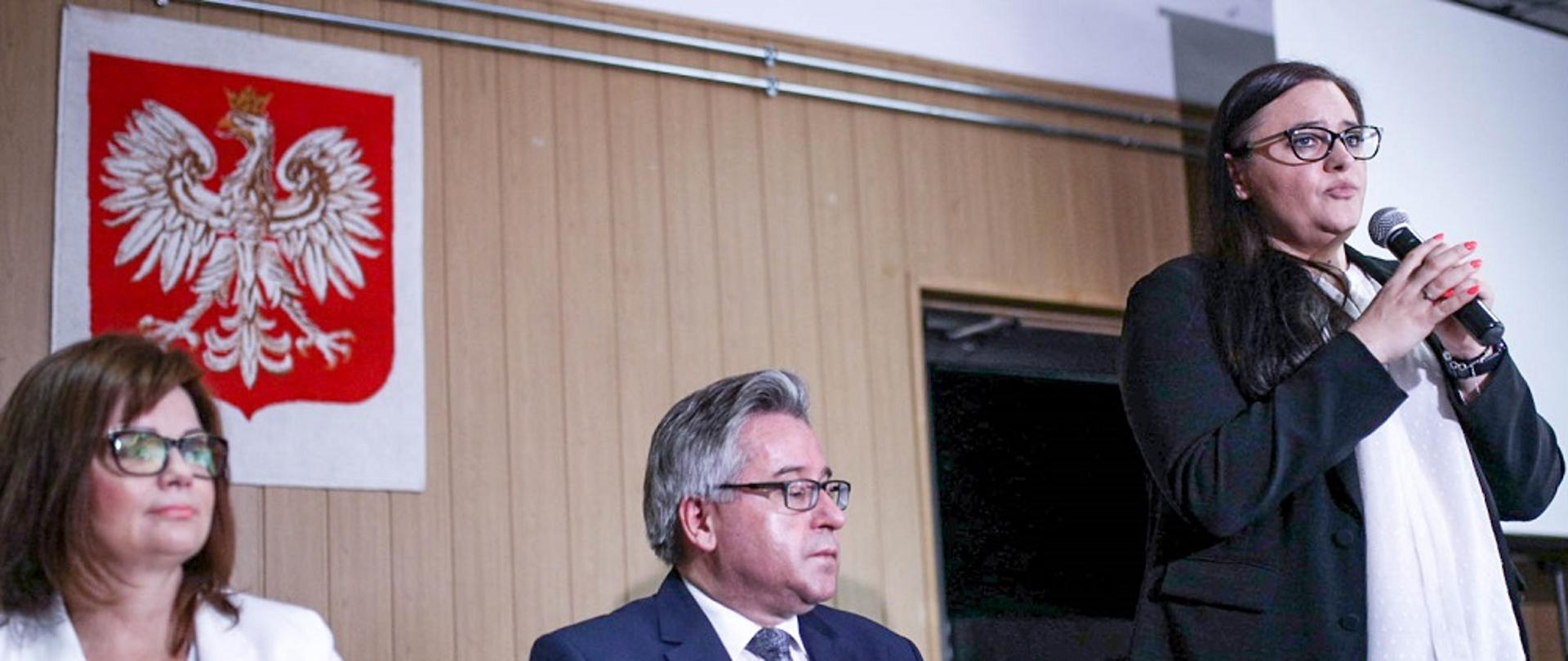 Na zdjęciu widoczne trzy osoby, mężczyzna i kobieta siedzą za stołem konferencyjnym. Minister Małgorzata Jarosińska-Jedynak stoi i mówi do mikrofonu