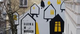 Mural przedstawiający stylizowane domy w kolorach czerni, bieli i żółci. Kompozycja przypomina wzgórze z unoszącymi się domkami o geometrycznych kształtach i żółtych oknach. Na jednym z domów widnieje napis „Głową w murach”. Przed muralem znajdują się bezlistne drzewa i fragment podwórza miejskiego.