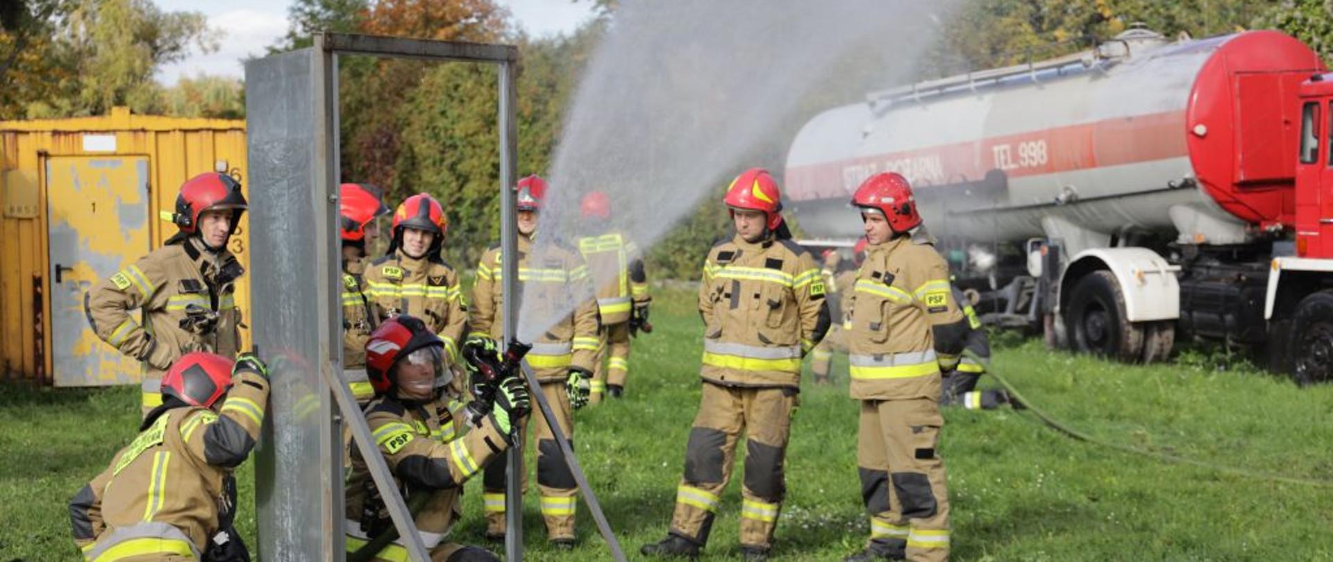 Strażacy PSP w umundurowaniu bojowym ćwiczą na poligonie KW PSP w Opolu posługiwanie się prądami gaśniczymi na różne obiekty i w różnych warunkach. W tle widoczne elementy poligonu strażackiego.