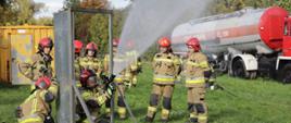 Strażacy PSP w umundurowaniu bojowym ćwiczą na poligonie KW PSP w Opolu posługiwanie się prądami gaśniczymi na różne obiekty i w różnych warunkach. W tle widoczne elementy poligonu strażackiego.