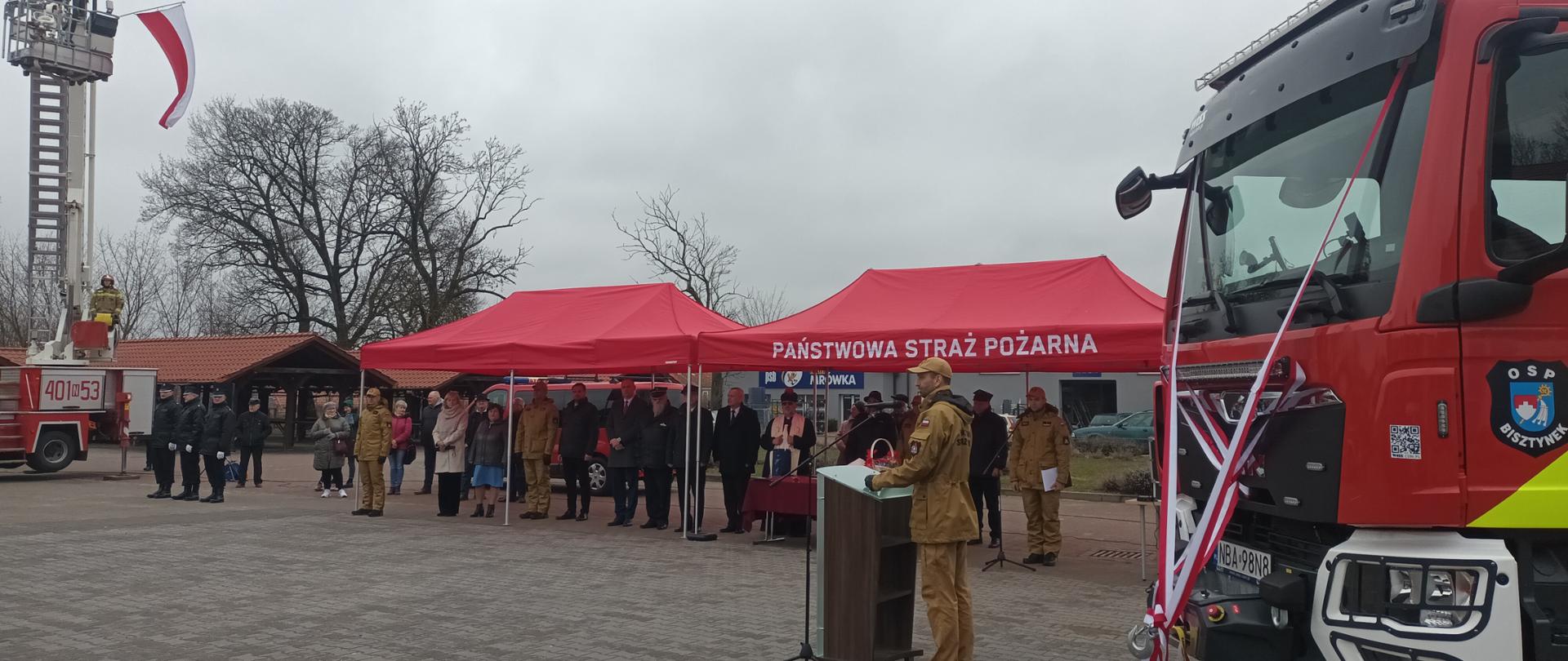 Przemowa Warmińsko - mazurskiego Komendanta Wojewódzkiego Państwowej Straży Pożarnej, po prawej stronie przód nowego pojazdu z oznaczeniami przynależności do OSP, w tle zaproszeni goście