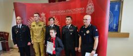 fotografia przedstawia laureata wraz z kadrą oraz egzaminatorami konkursu, w tle ścianka Państwowa Straż Pożarna, część wyposażenia Sali